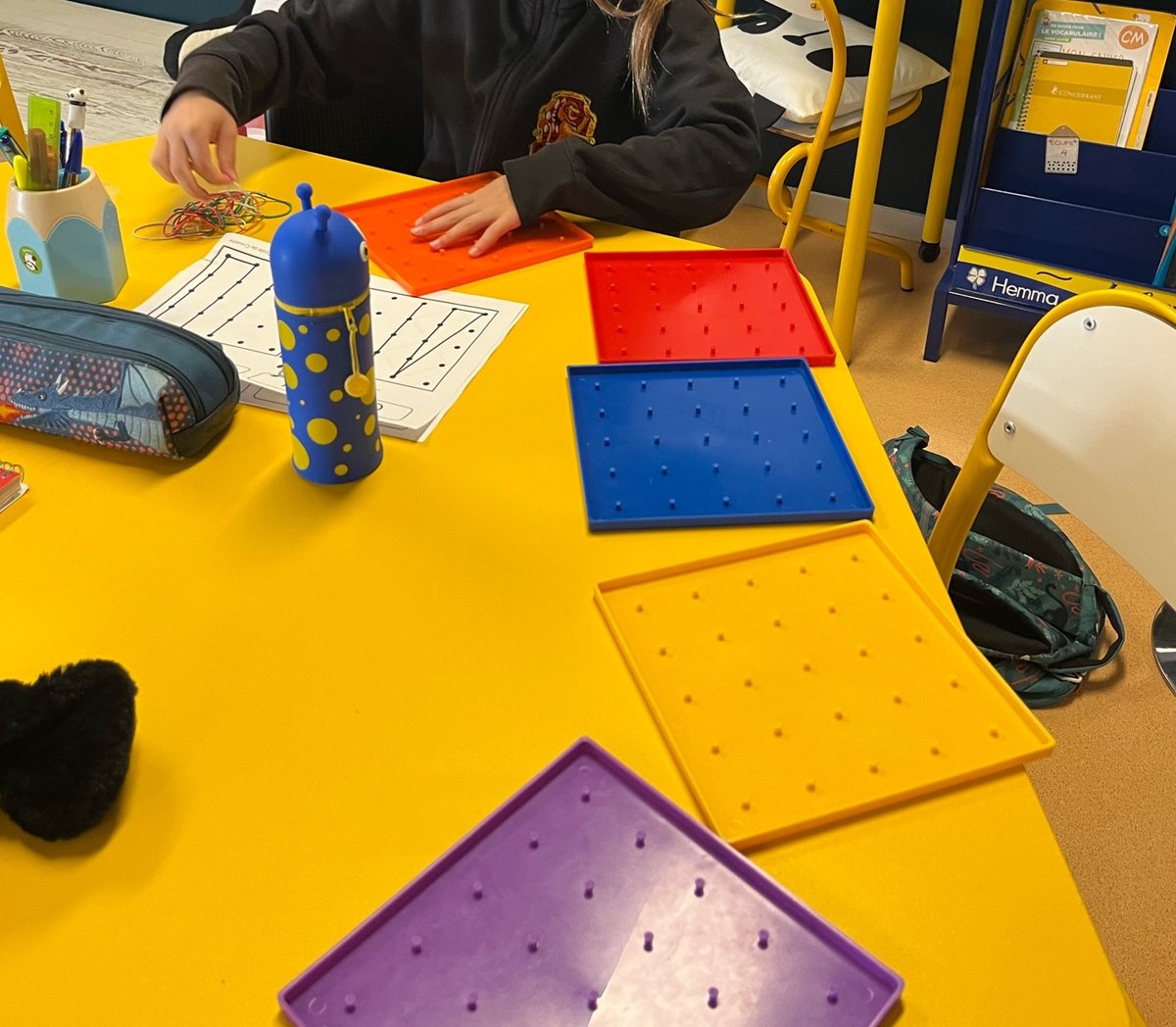 Géoplans en plastique de couleur posés sur une table jaune, élastiques et fiche de figures à reproduire