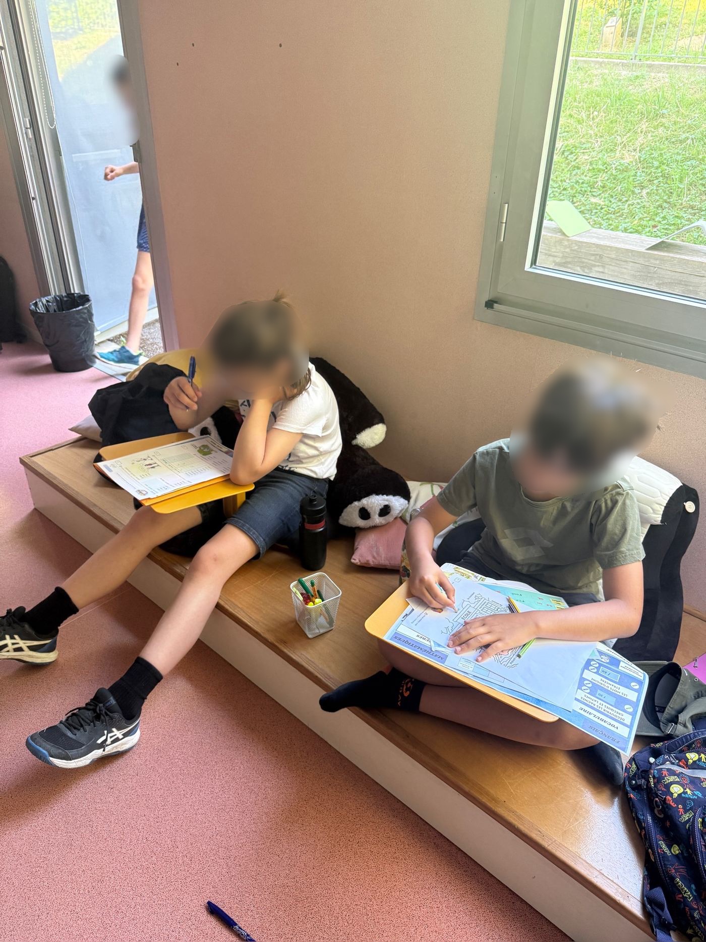 Deux élèves installés dans un coin lecture, l'une au sol avec un dossier, l'autre adossée à un grand panda en peluche, plongés dans leur travail.