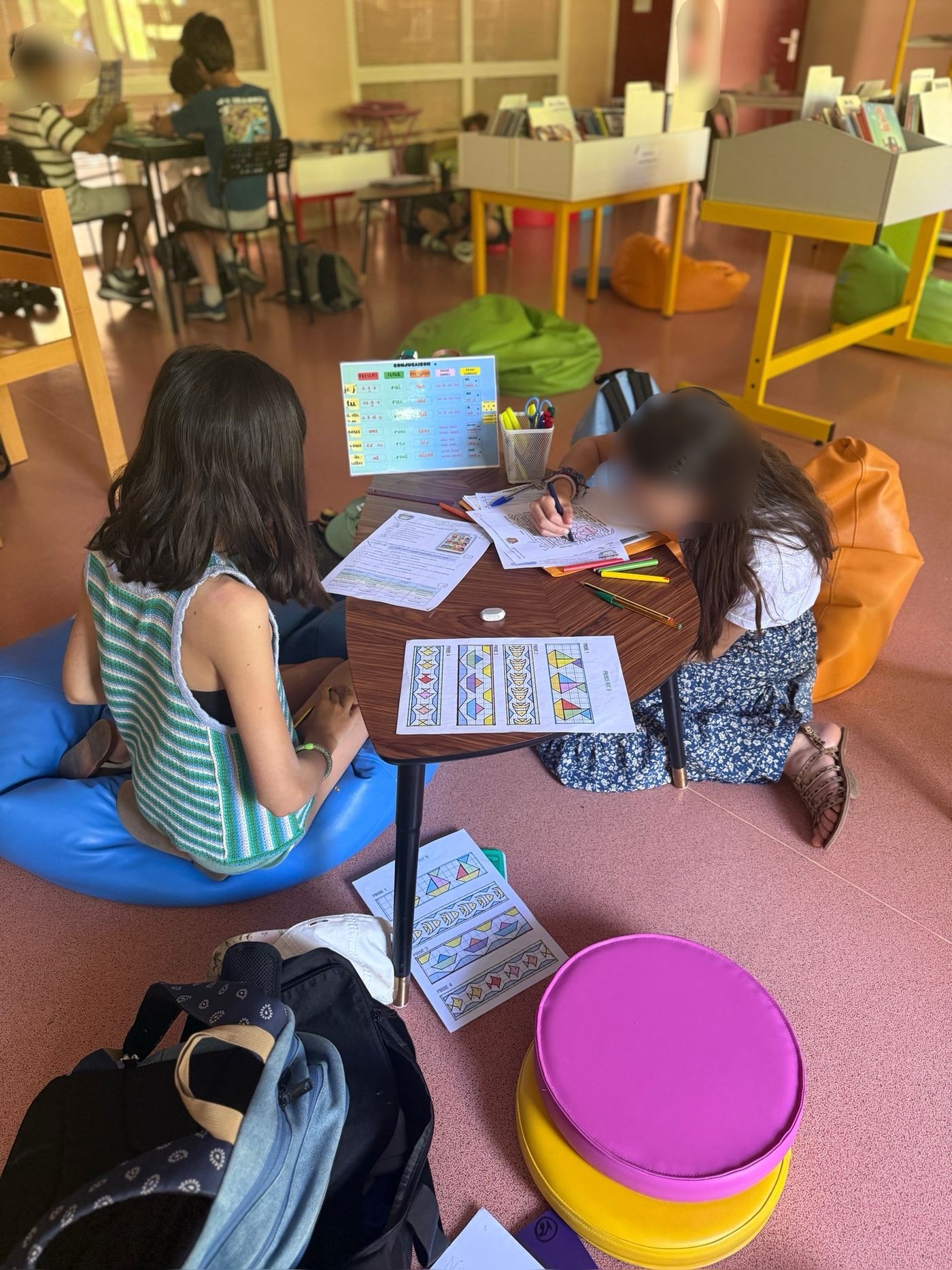 Élèves au travail dans la bibliothèque, sur des poufs colorés, autour d'une petite table basse en bois.